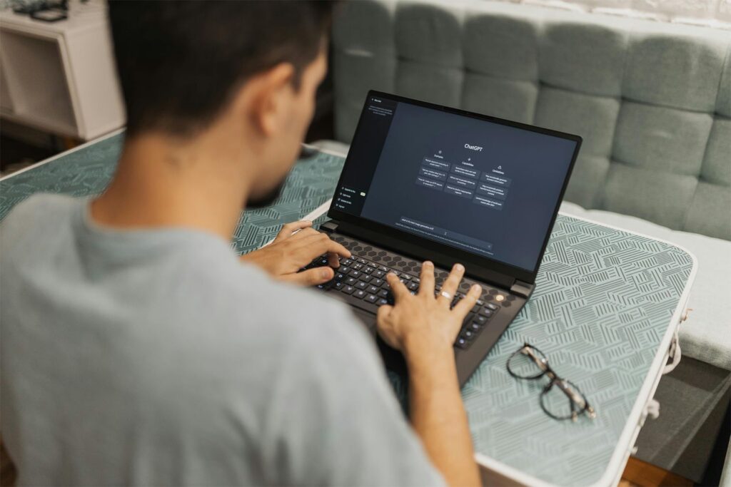 A person types on a laptop displaying the ChatGPT interface while sitting at a patterned table, possibly composing Claude AI email prompts, with eyeglasses resting nearby. The background shows a light-colored cushioned bench.