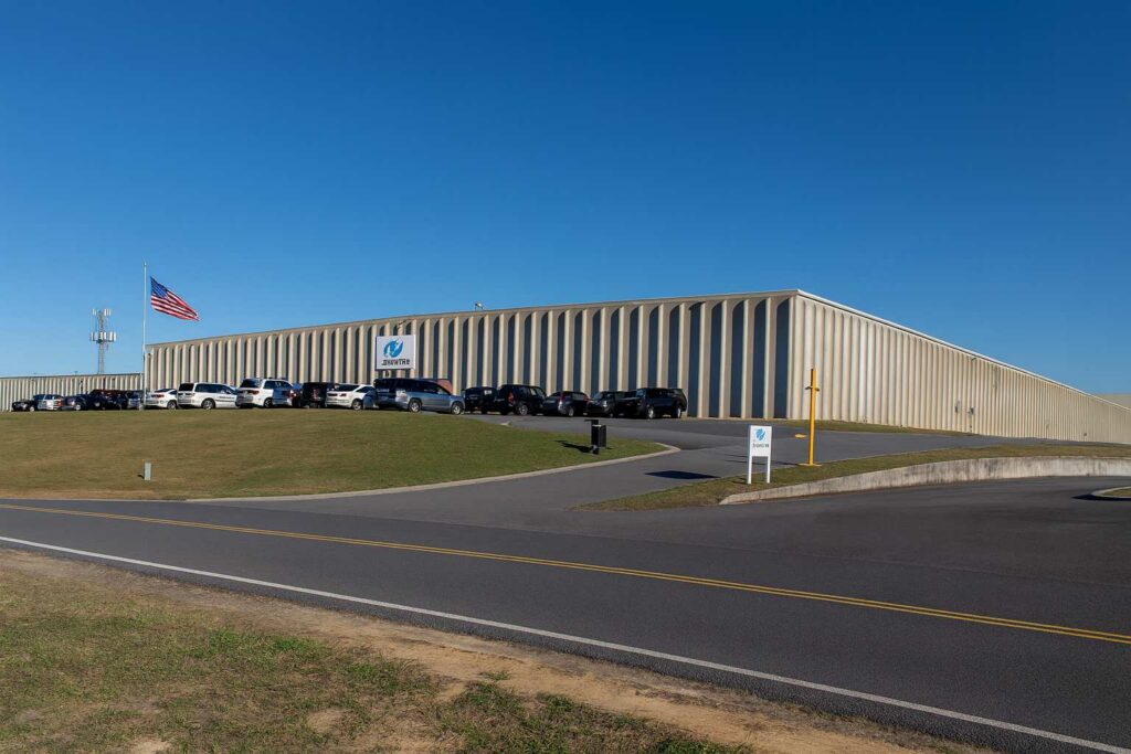 A large, beige industrial building with vertical siding sits behind parked cars and a U.S. flag under a clear blue sky. A sign and driveway are visible in the foreground, suggesting Kobayashi's expertise in product development workflow software.