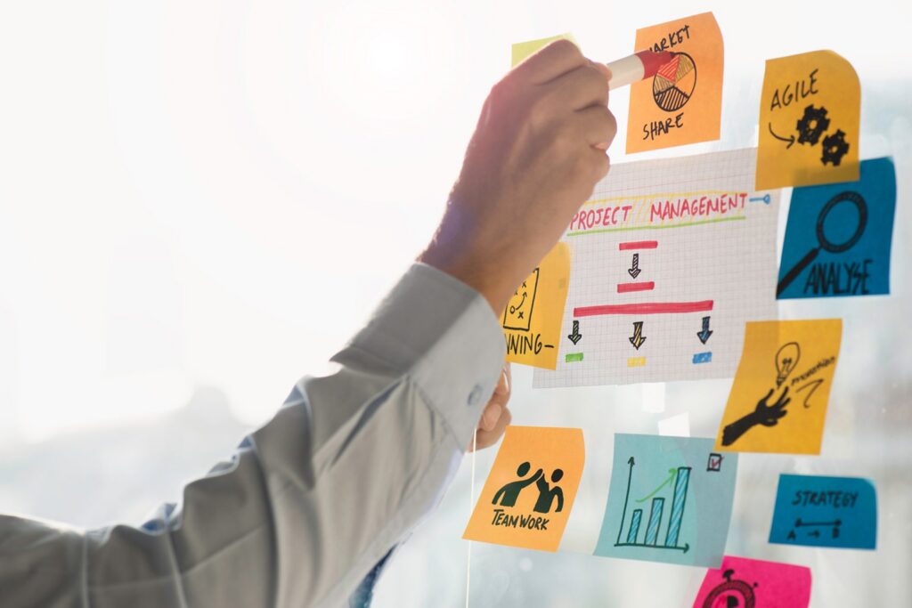 A person points to a project management board with colorful sticky notes displaying charts, graphs, and words like AGILE and TEAMWORK, highlighting planning, organization, and project accountability in successful teams.