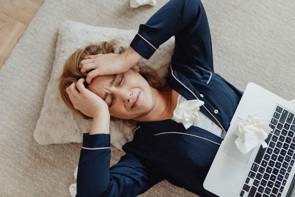 A person in dark pajamas lies on a carpet, clutching their head in frustration or pain, surrounded by crumpled tissues and a laptop—perhaps overwhelmed by workload planning or struggling to manage team workload.