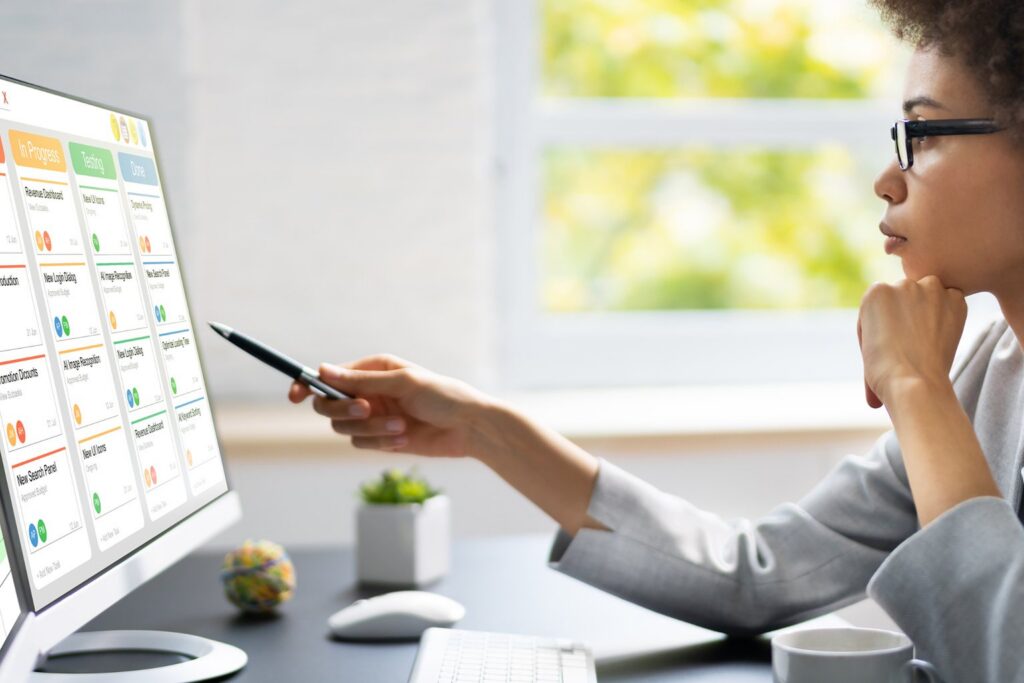 A person sits at a desk, looking intently at a computer monitor displaying a colorful project management dashboard featuring some of the best monday.com alternatives. They hold a pen and appear to be pointing at or interacting with the screen.