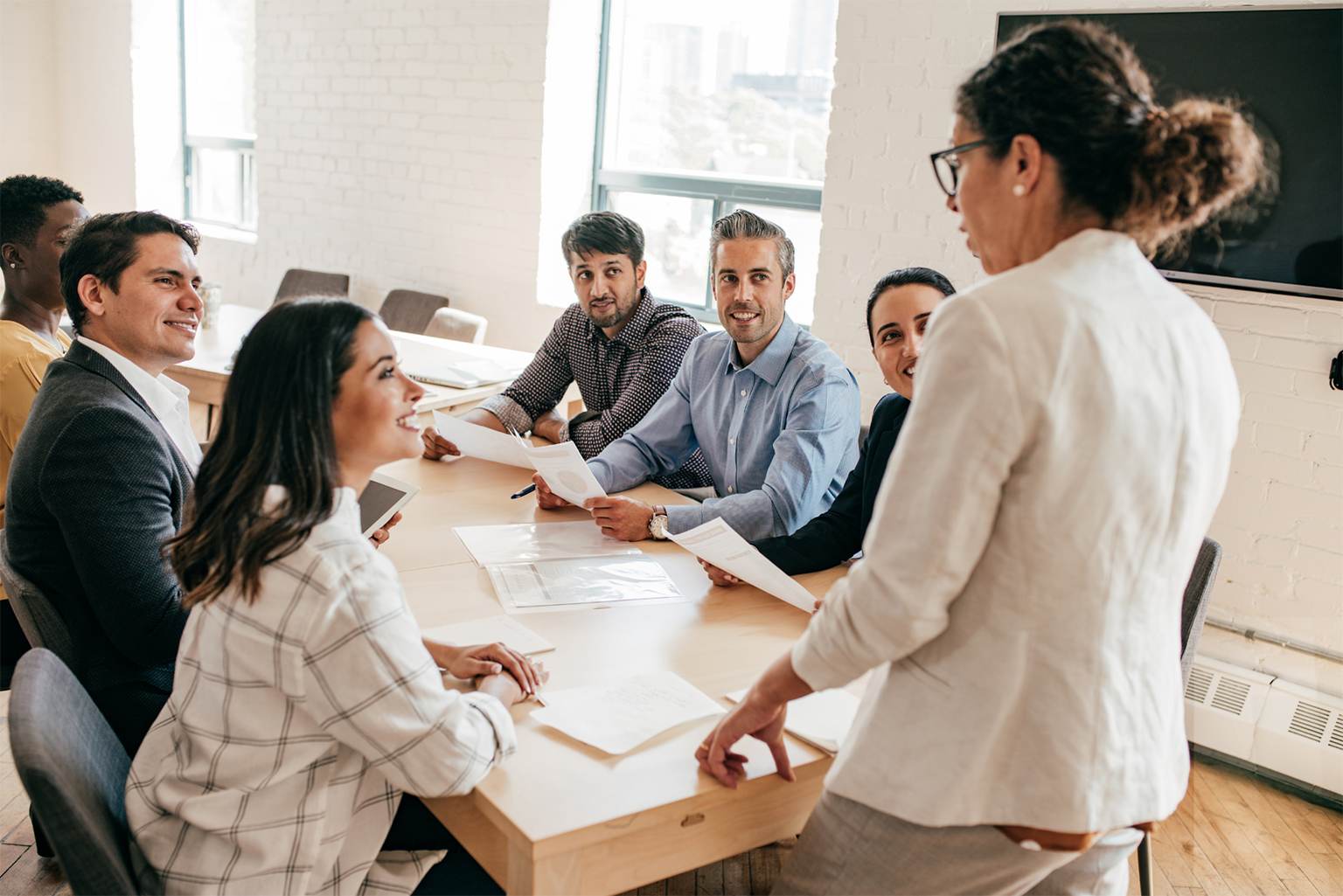 A group of six people sit around a table in a bright office, holding papers and listening attentively to a woman presenting on Microsoft Project Online 2026. A large TV screen is mounted on the wall behind her.