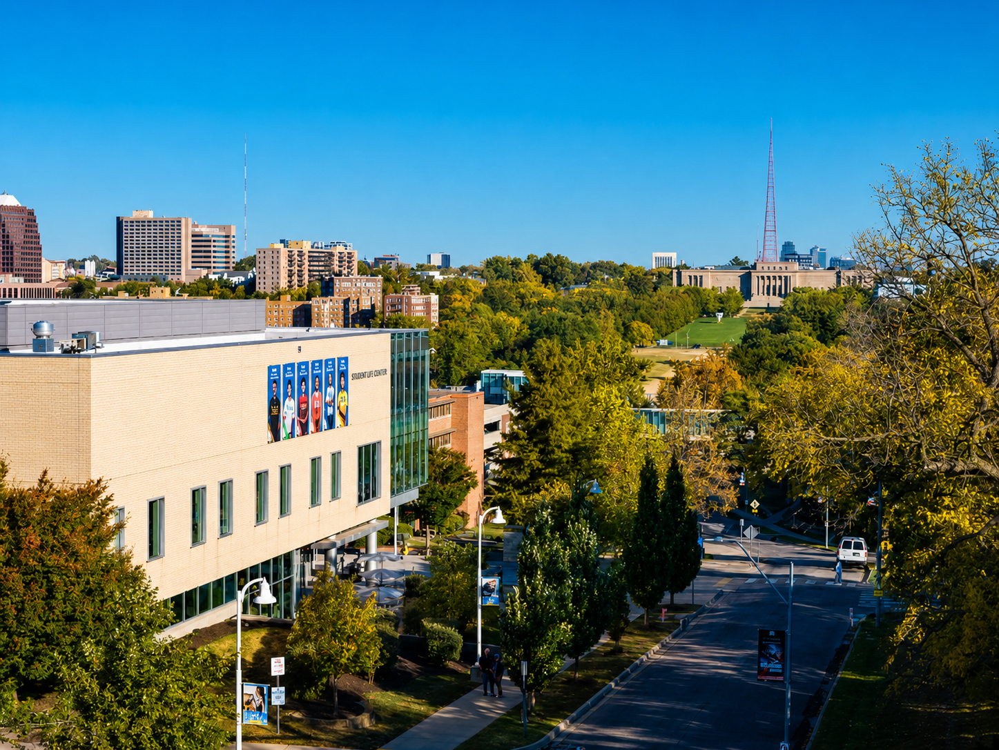 An aerial view of a city campus with a modern building on the left, tree-lined streets, and a distant tower and large classical building under a clear blue sky—an inspiring setting for mastering project management workflow.