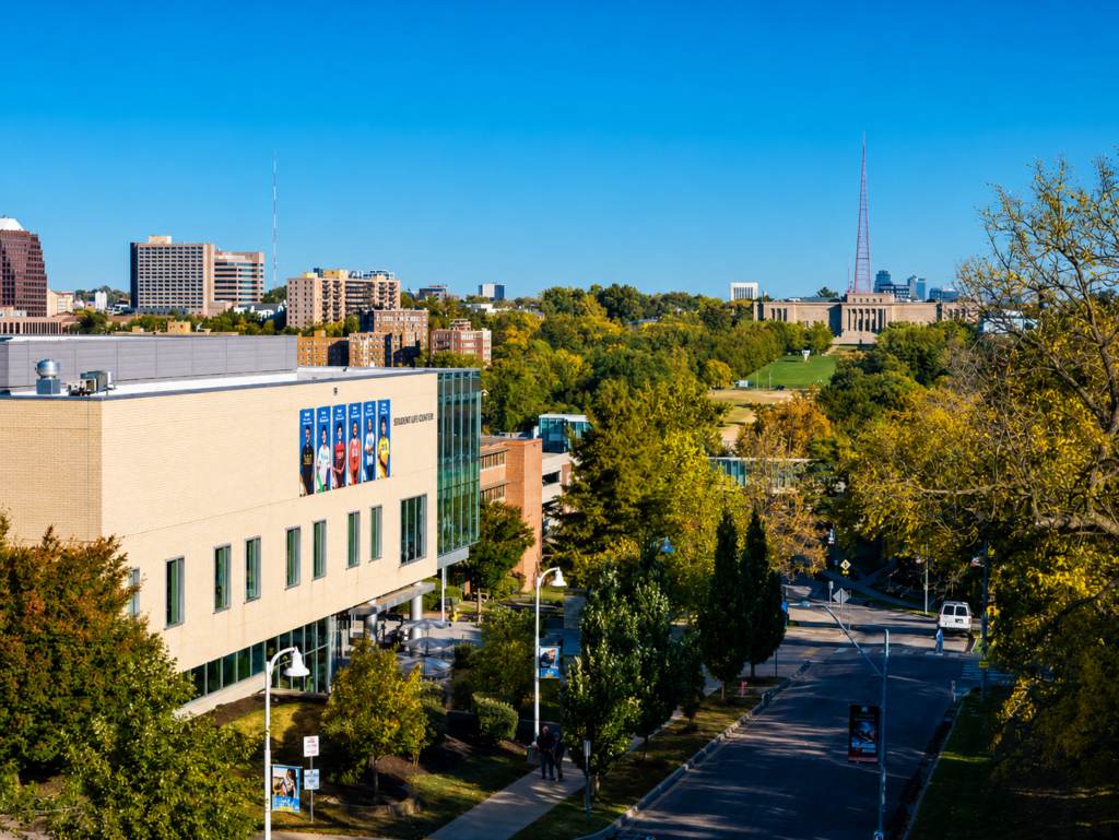 An aerial view of a city campus with a modern building on the left, tree-lined streets, and a distant tower and large classical building under a clear blue sky—an inspiring setting for mastering project management workflow.