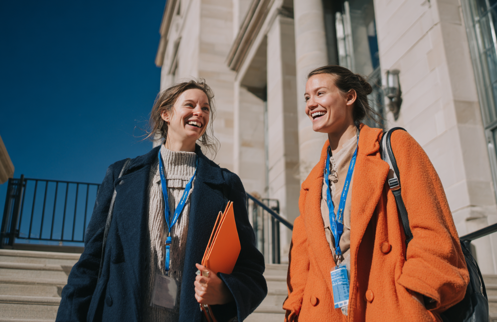 Two smiling women wearing lanyards and coats, one in blue and one in orange, walk down steps outside a large stone building on a sunny day, holding folders related to Higher Education IT and looking at each other.